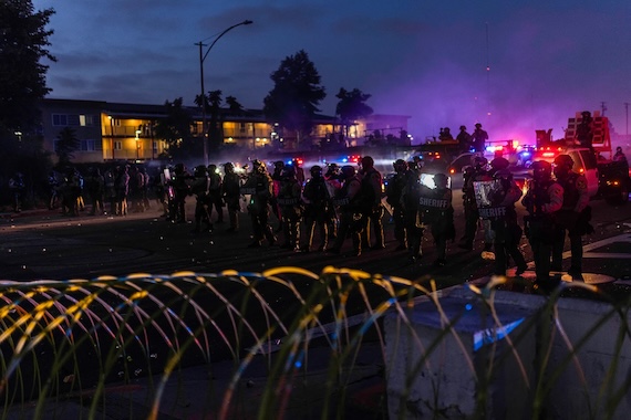 A line of sheriff’s deputies in riot gear and tactical vests stand in formation at night during a protest. Police vehicles with flashing lights illuminate the scene, casting red and blue hues across the street. In the foreground, coiled razor wire partially obstructs the view, and in the background, purple smoke drifts through the air in front of apartment buildings with lit windows.